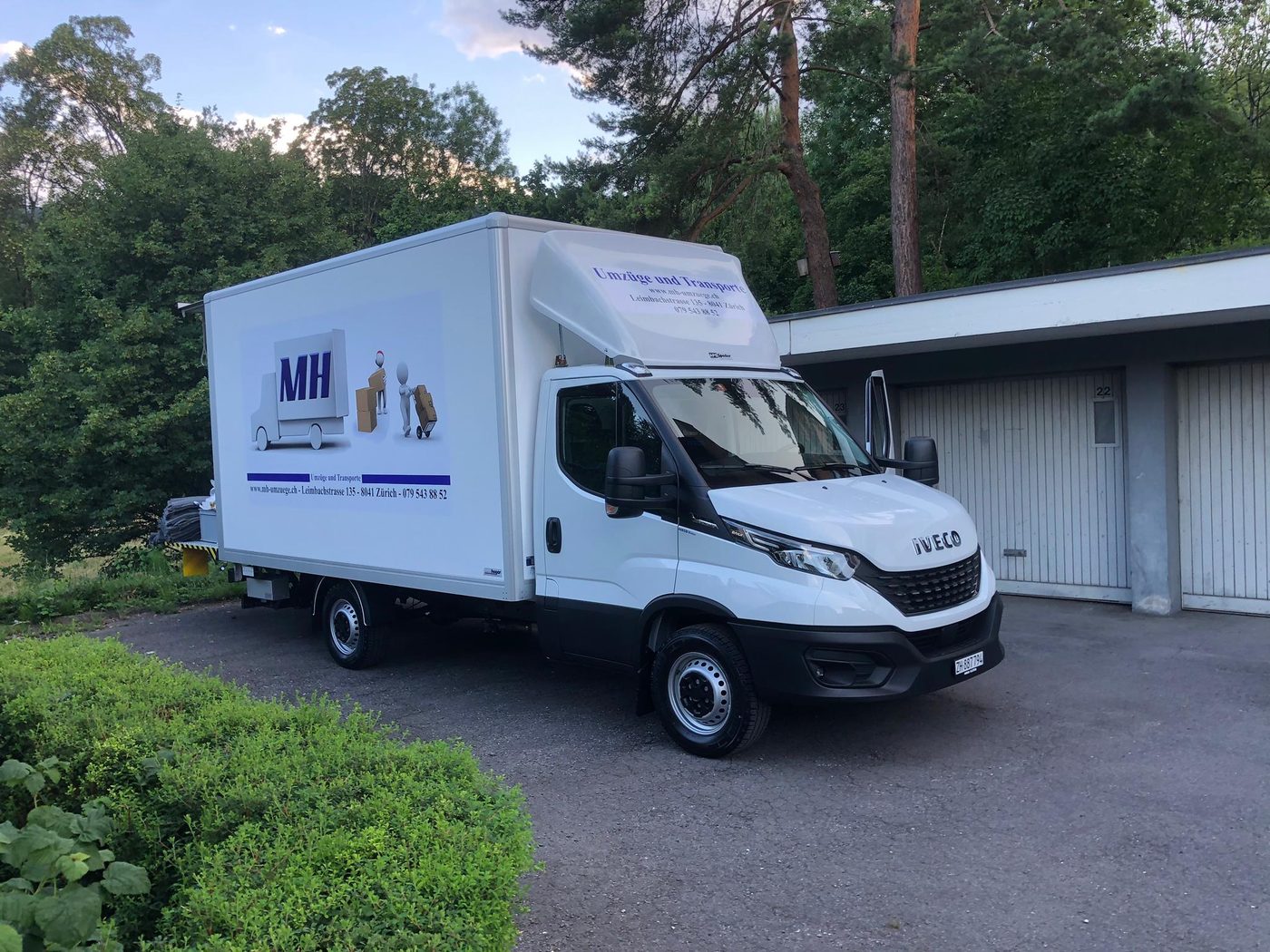 Weisser MH-Lastwagen mit Firmenlogo auf einem Kiesplatz vor Garagen